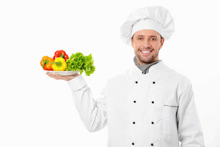 Young cook holding a plate with vegetables on white backgroundの写真素材