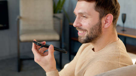 Man sitting on sofa holds phone talks on speakerphone, record audio message indoorsの写真素材