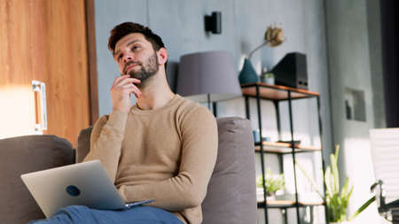 Young man looking at the ceiling with laptop sitting on a couch at homeの写真素材