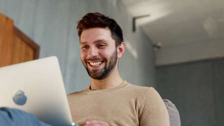 Young smiling man communicating by videoconference in his apartment. Happy man with laptop in...