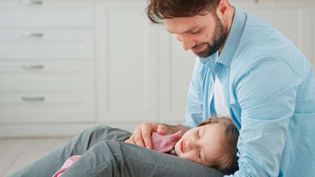 Smiling dad with beard looks and caresses small girl child with long fair hair sleeping on father knees at home close viewの写真素材
