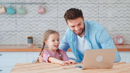 Smiling family with laptop in the kitchen. Young dad and little daughter talking on an online conference with mom using a laptopの写真素材