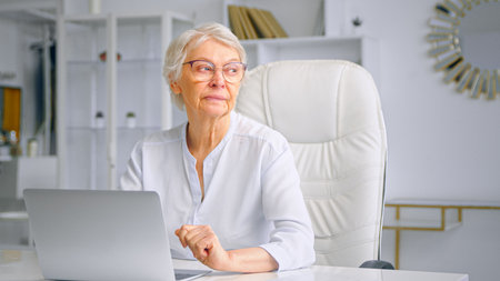 Concentrated aged lady manager in glasses sitting on large chair at table against home furniture