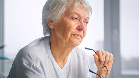 Serious senior businesswoman with short gray hair holds glasses in trembling hand against blurred...