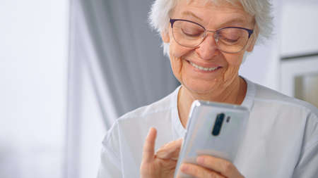 Happy aged in glasses and blue blouse communicates with children on white smartphone against blurred...