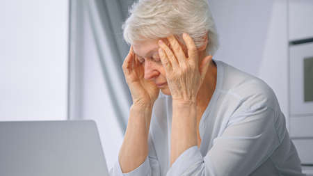 Old tired of work lady pensioner with short gray hair massages temples with hands sitting by laptop at home close viewの写真素材