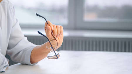 Thoughtful old businesswoman holds glasses over white table against blurred large home window close viewの写真素材