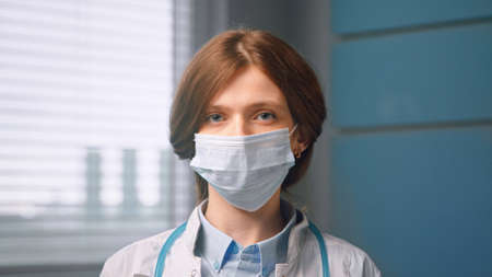 Red haired woman with face mask and looks straight standing in local hospital room close viewの写真素材