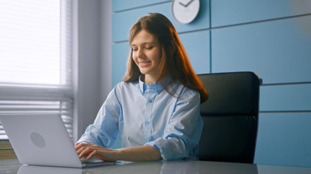 Young woman general practitioner communicates with patients online typing on gray laptop and smiling at table in doctor officeの写真素材
