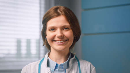 Pretty cheerful young woman general practitioner looks straight and smiles standing against blue hospital room wall close viewの写真素材