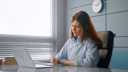 Concentrated young woman doctor writes in paper notebook listening to speaker at online medical conference via laptop in officeの写真素材