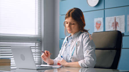 Young woman therapist in white coat talks to patient holding papers sitting on chair by wall with body pictures at office tableの写真素材