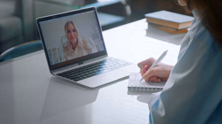 Medical university lady student in blue coat writes lecture in notebook sitting at online lesson via laptop with lecturer closeupの写真素材