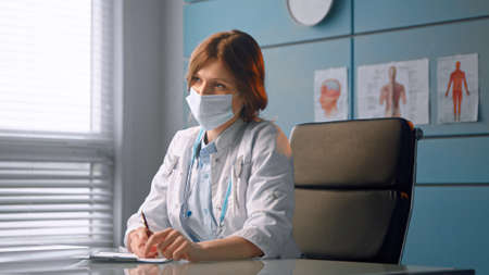 Young woman doctor in white medical coat talks to new patient and writes patient record before examination in local hospitalの写真素材