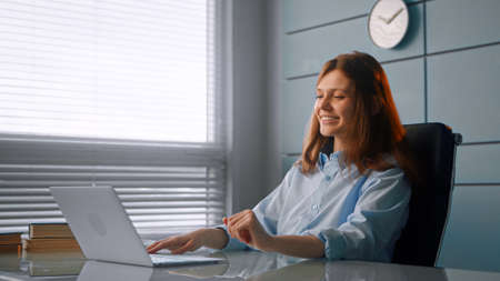 Smiling worker brunette with long loose hair types on gray laptop and smiles cheerfully sitting at table in officeの写真素材