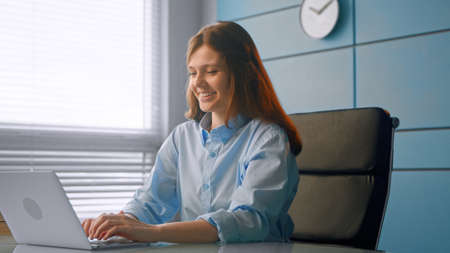 Smiling woman general practitioner communicates with patients online typing on gray laptop and smiling at table in doctor officeの写真素材