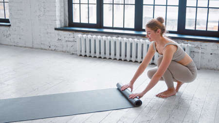 Slim blonde barefoot sportswoman in light beige tracksuit rolls rubber mat after training near large window in spacious yoga studioの写真素材