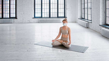 Tranquil young woman in tracksuit and protective mask meditates sitting in lotus pose in spacious studio with large windowsの写真素材