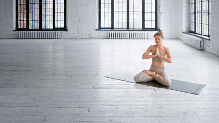 Athletic barefoot lady in tracksuit sits on mat in lotus pose with namaste mudra meditating in spacious studio with windowsの写真素材