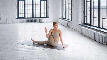 Flexible blonde lady in stylish light gray tracksuit does half lord of fishes pose sitting on mat in studio with large windowsの写真素材