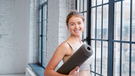 Blonde woman fitness trainer with hair bun holding mat looks out of window and turns to camera in modern yoga studio closeupの写真素材