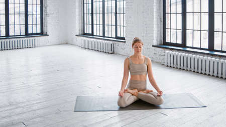Peaceful young woman in stylish tracksuit sits on mat in full lotus yoga pose meditating in spacious gymの写真素材