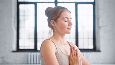 Pretty woman with closed eyes in beige sportswear breathes deeply holding hands in namaste mudra at chest in yoga studio closeupの写真素材