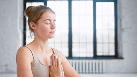 Blonde lady with closed eyes in beige top breathes deeply holding hands in namaste mudra by chest in light yoga studio closeupの写真素材