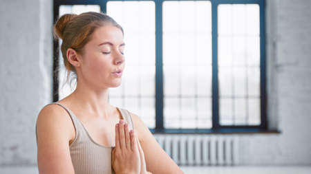 Calm young woman with hair bun in beige top does yoga breathing exercises holding hands in namaste mudra in studio close viewの写真素材
