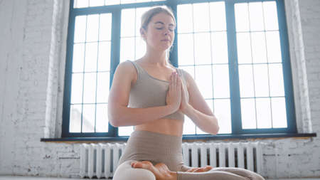 Concentrated young woman in stylish beige tracksuit does padma asana with namaste mudra meditating in spacious yoga studioの写真素材