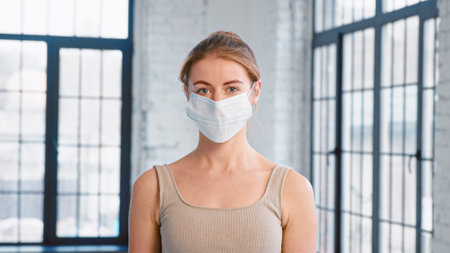 Young lady trainer in tracksuit takes off protective mask looking into camera in spacious studio with windows closeupの写真素材