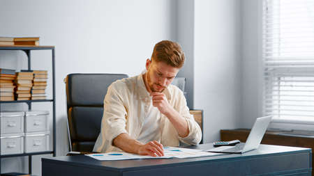 Bearded male financial director reads bank reports sitting at gray table and calculator in office near large windowの写真素材