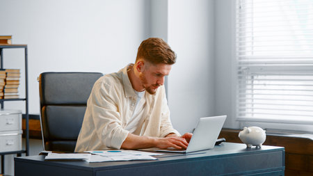 Concentrated businessman signs contract online and raises hands sitting near white laptop at gray office tableの写真素材