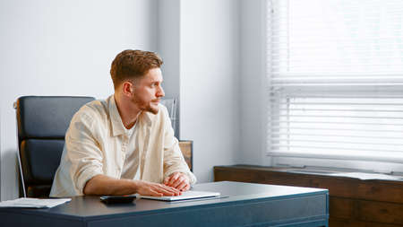 Thoughtful guy manager with beard sums up administrative expenses on calculator and thinks on reducing costs at table in officeの写真素材