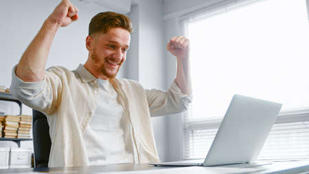 Concentrated businessman signs contract online and raises hands with happiness sitting near white laptop at gray office tableの写真素材