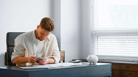 Serious bearded man accountant counts dollar banknotes checking with paper reports on gray table with piggy bank in new officeの写真素材