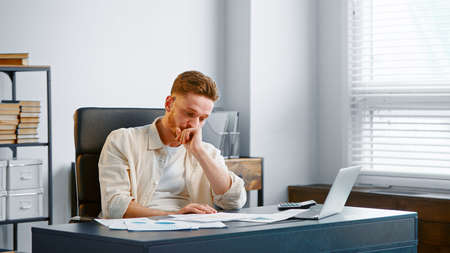 Upset man reads bank reports sitting at gray table and calculator in officeの写真素材