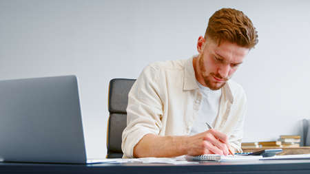 Startup company accountant in yellow shirt summarizes unexpected expenses on calculator and writes into paper notebook at homeの写真素材