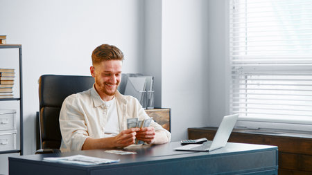 Successful businessman with beard counts cash money with happy smile sitting at table with white laptop against office windowの写真素材