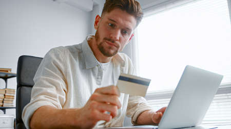 Company freelance bearded manager types yellow card number on white laptop conducting online payment at table staying homeの写真素材
