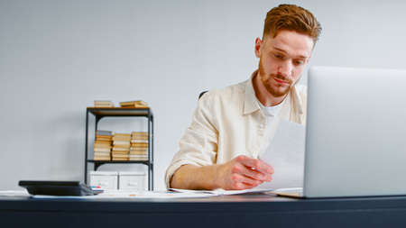 Concentrated financial director with beard looks through papers with business project at table with laptop and calculator in officeの写真素材