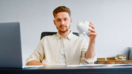 Serious man corporate company manager with beard holds white piggy bank and smiles joyfully posing at office table with laptopの写真素材