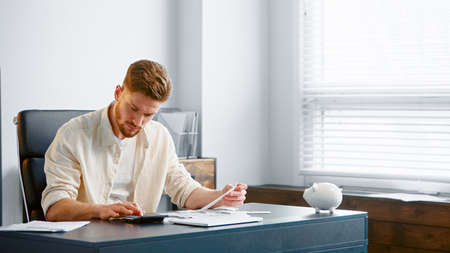 Thoughtful guy manager with beard sums up administrative expenses on calculator and thinks on reducing costs at table in officeの写真素材