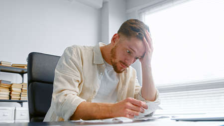 Shocked man bank accountant with beard looks at paper checks and puts hand on head after sorting receipts on table in officeの写真素材