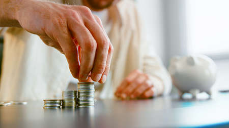 Careful guy company manager puts saved silver coins on stack on gray office table against blurred piggy bank at home closeupの写真素材