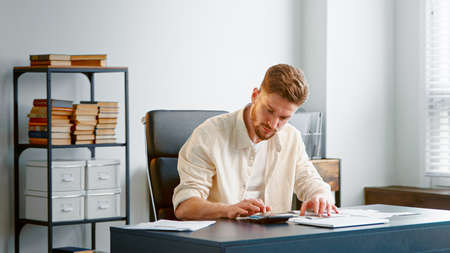 Professional accountant with beard counts paper checks summarizing sums on calculator with serious face sitting at table in officeの写真素材