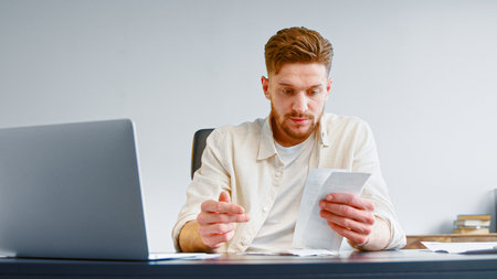 Bearded professional accountant sorts paper invoice receipts sitting at table with white laptop in company office closeupの写真素材