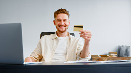 Smiling guy successful company manager with beard holds credit card and poses sitting at gray table with laptop and papersの写真素材