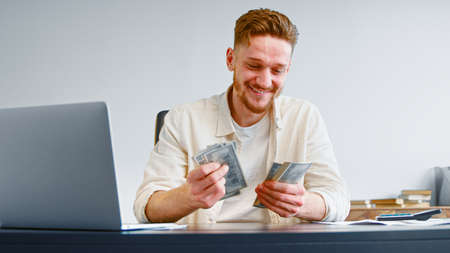 Smiling self employed businessman counts cash income holding dollar banknotes sitting at table with white laptop and calculatorの写真素材