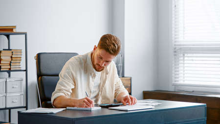 Bearded accountant sums up unexpected expenses from checks on calculator and writes results in paper notebook sitting in officeの写真素材